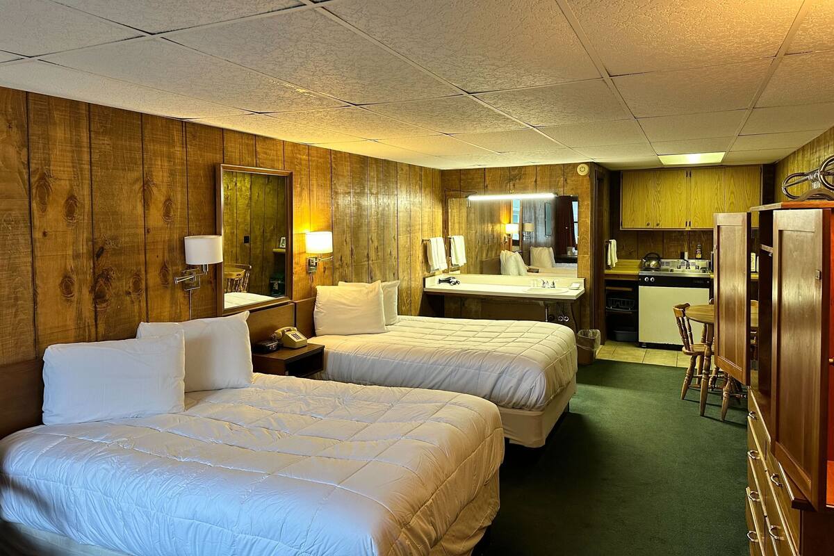 Quarto de hotel com duas camas, banheiro, cozinha compacta e parede de madeira.