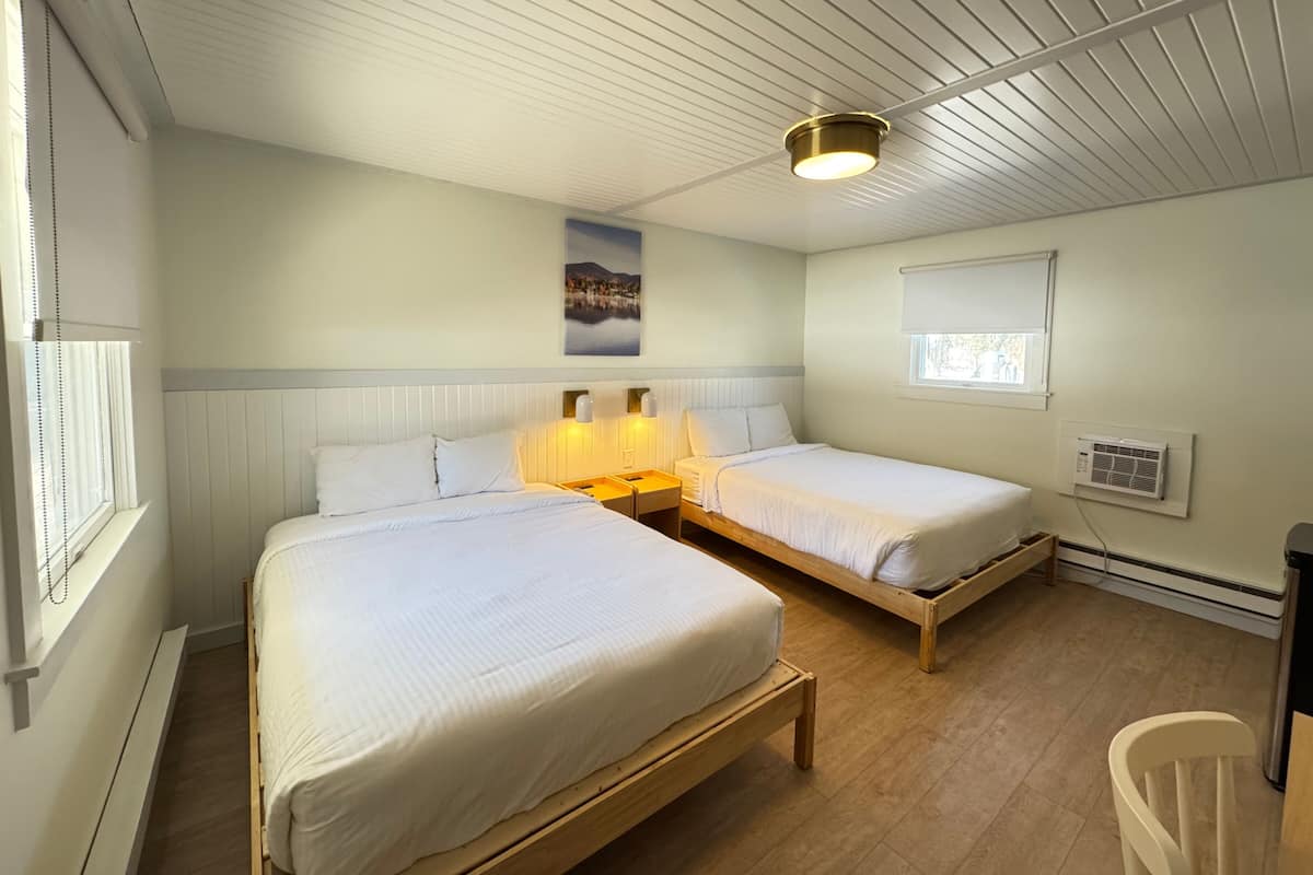 Quarto com duas camas, uma luminária no teto, janela com persianas e ar-condicionado fixado na parede.