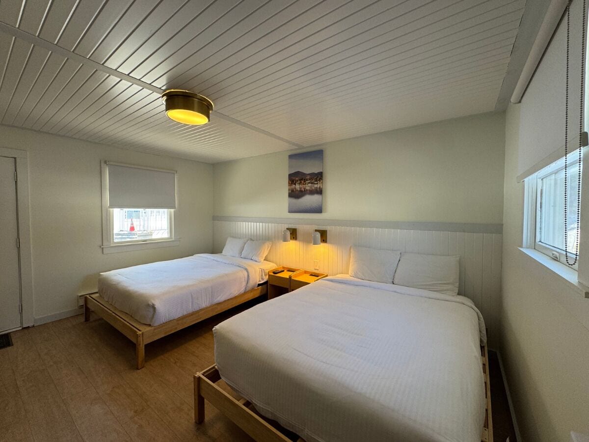 Et værelse med to senge, trægulv, loft med vandrette linjer, et vindue med persienner og en lampe på væggen.