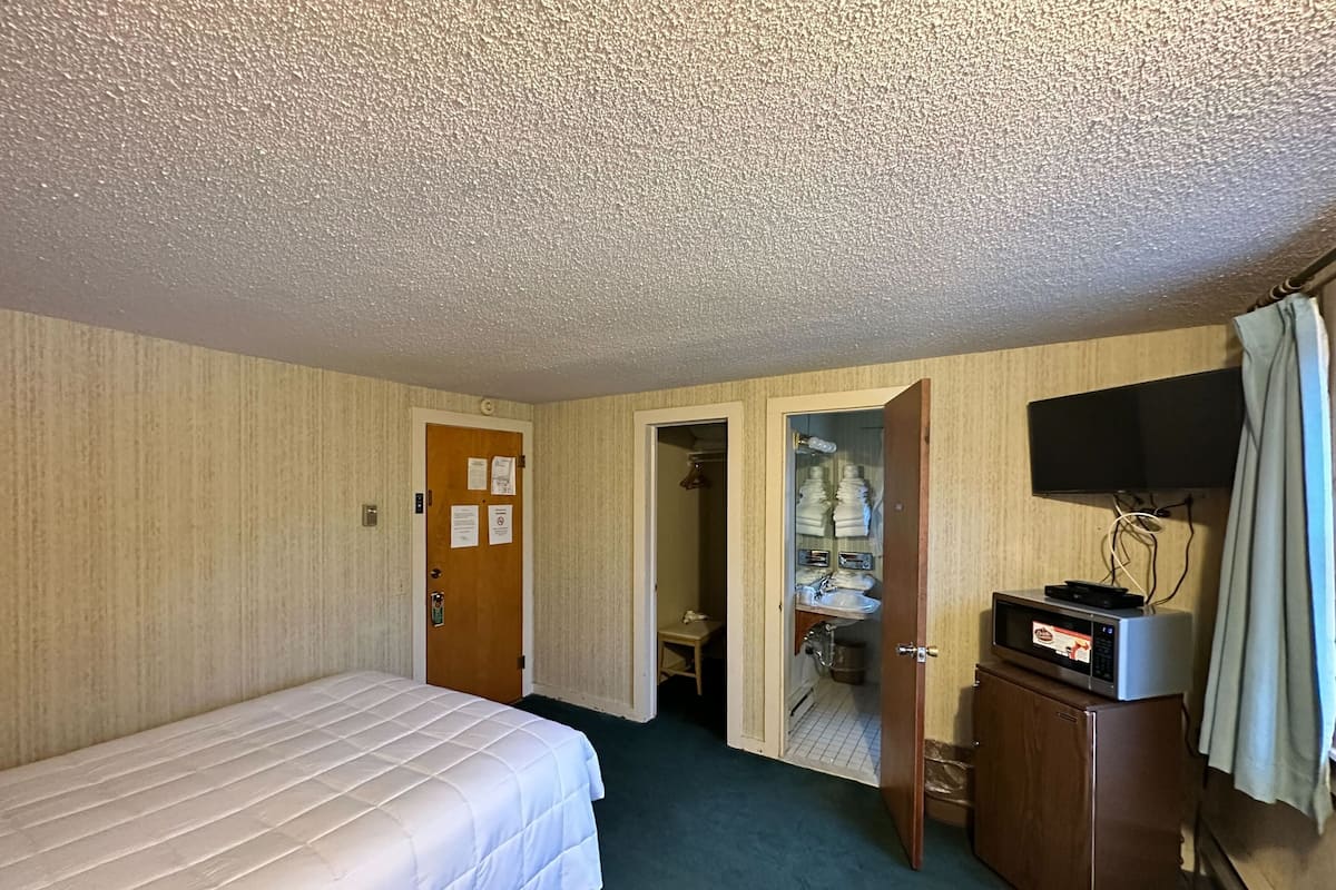 Quarto de hotel com cama, escrivaninha com televisão, banheiro com pia e porta-toalhas, e porta com avisos.