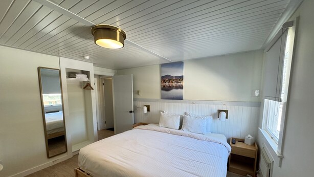 A hotel room with a large bed, a bedside table, a nightstand, a mirror, a wardrobe, and a window with a view of a lake and mountains. - Maple Leaf Inn - Lake Placid (Lake Placid)