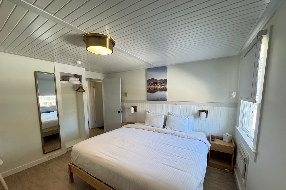 Quarto de hotel com cama grande, mesa de cabeceira, espelho, guarda-roupa e janela com vista para um lago e montanhas.