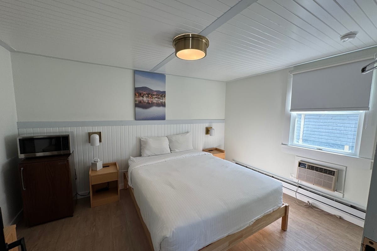 Quarto com cama, mesa de cabeceira, escrivaninha, micro-ondas e janela com persianas.