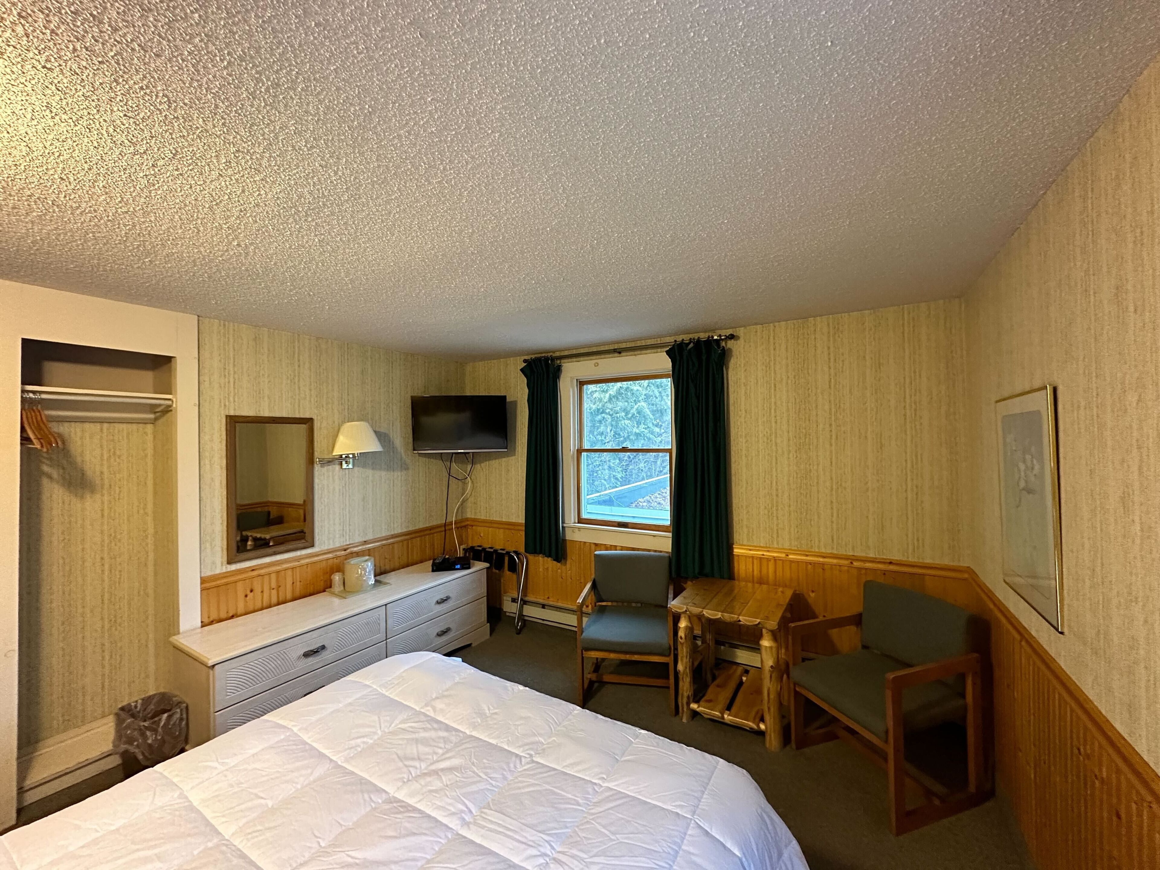 A hotel room with a bed, a desk, a chair, a TV, a window with curtains, and a mirror.
