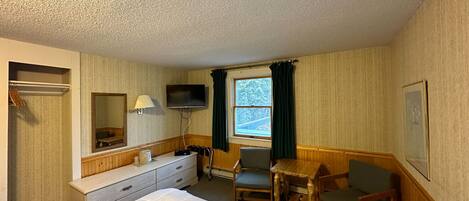 Quarto de hotel com cama, escrivaninha, cadeira, televisão, janela com cortinas e espelho.