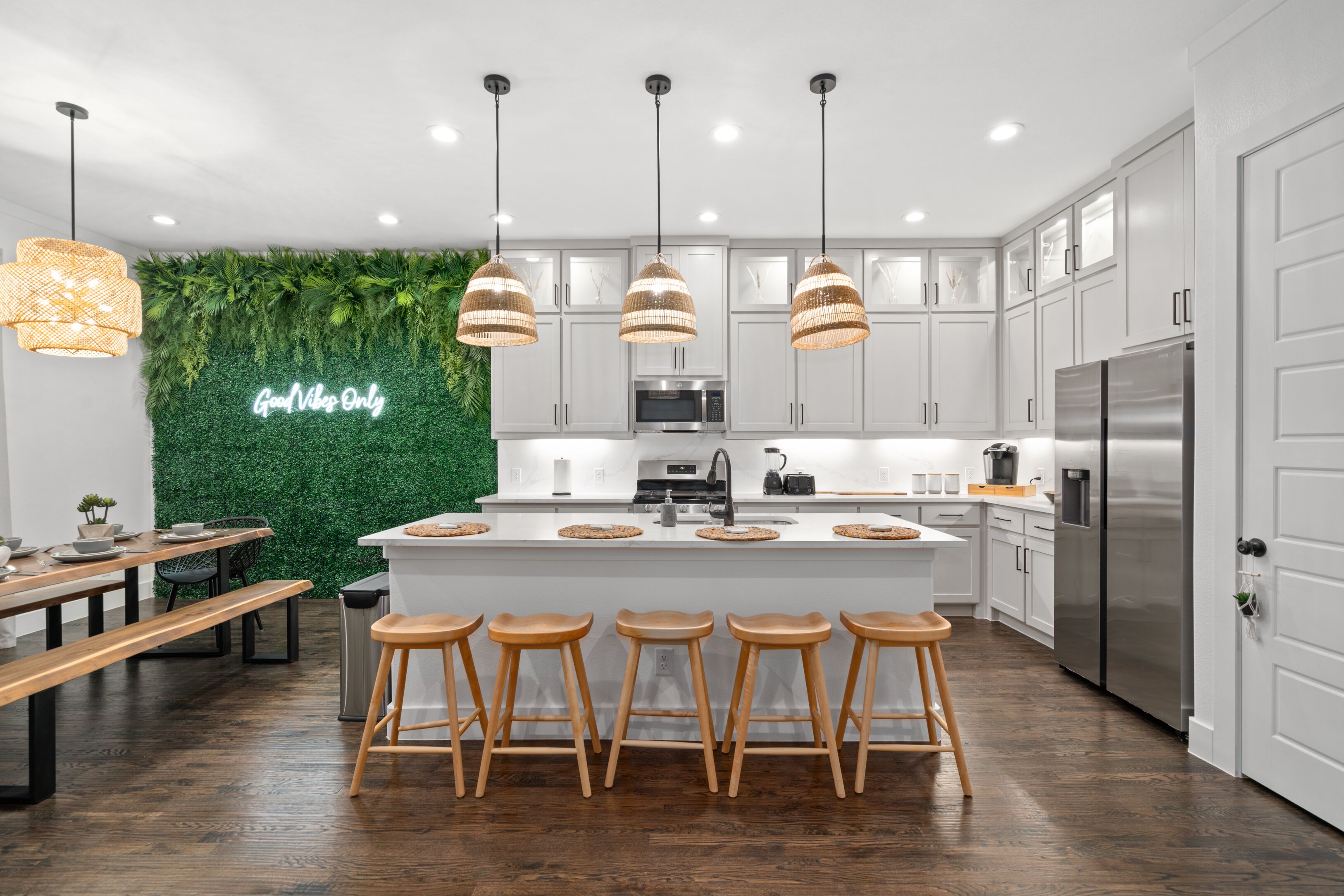 Una cocina moderna con armarios blancos, una isla central con taburetes, una pared verde con un letrero de neón y lámparas colgantes.