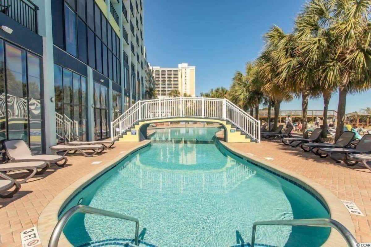 A swimming pool with lounge chairs and a bridge, surrounded by palm trees and buildings.
