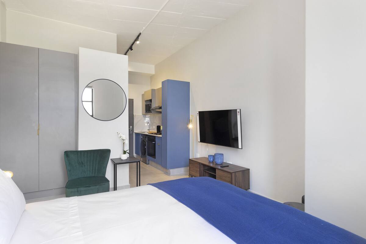 Habitación de hotel con cama, una silla verde, una mesita pequeña, televisor y una kitchenette al fondo.