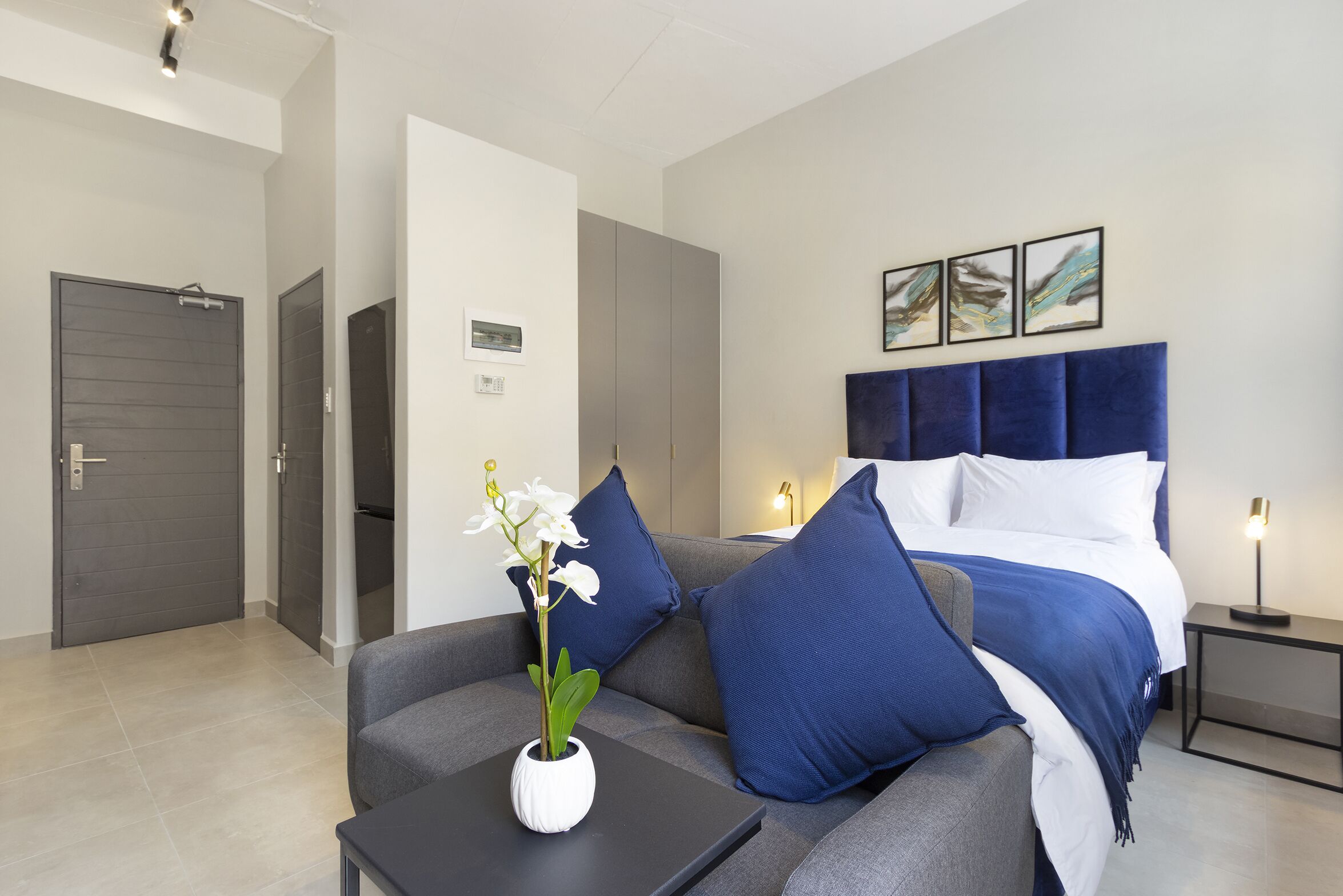 Une chambre d’hôtel moderne avec un canapé gris, un vase blanc orné de fleurs, un lit avec une literie bleue et une table de chevet avec une lampe.