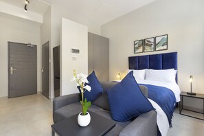 Quarto de hotel moderno com um sofá cinzento, um vaso branco com flores, uma cama com roupa de cama azul e uma mesa de cabeceira com uma luminária.