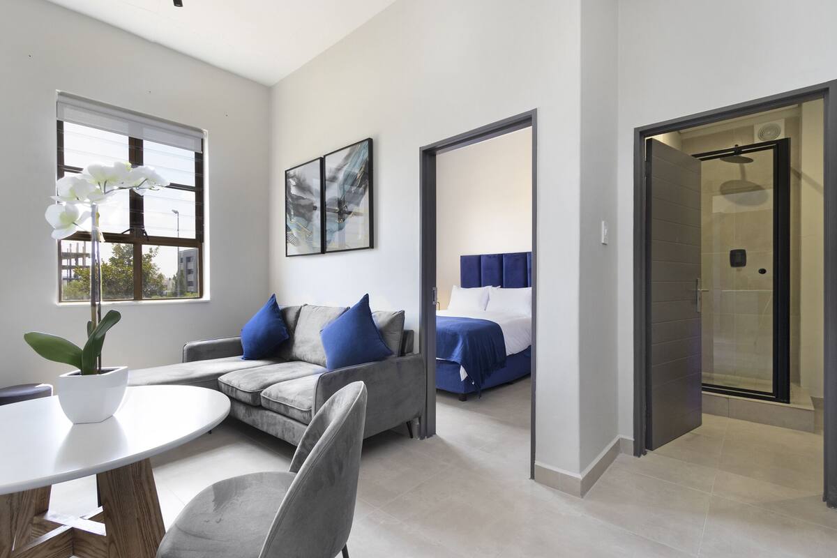 Una habitación de hotel moderna con un sofá gris, una cama con ropa de cama azul, una planta de orquídea blanca y un cuadro en la pared.