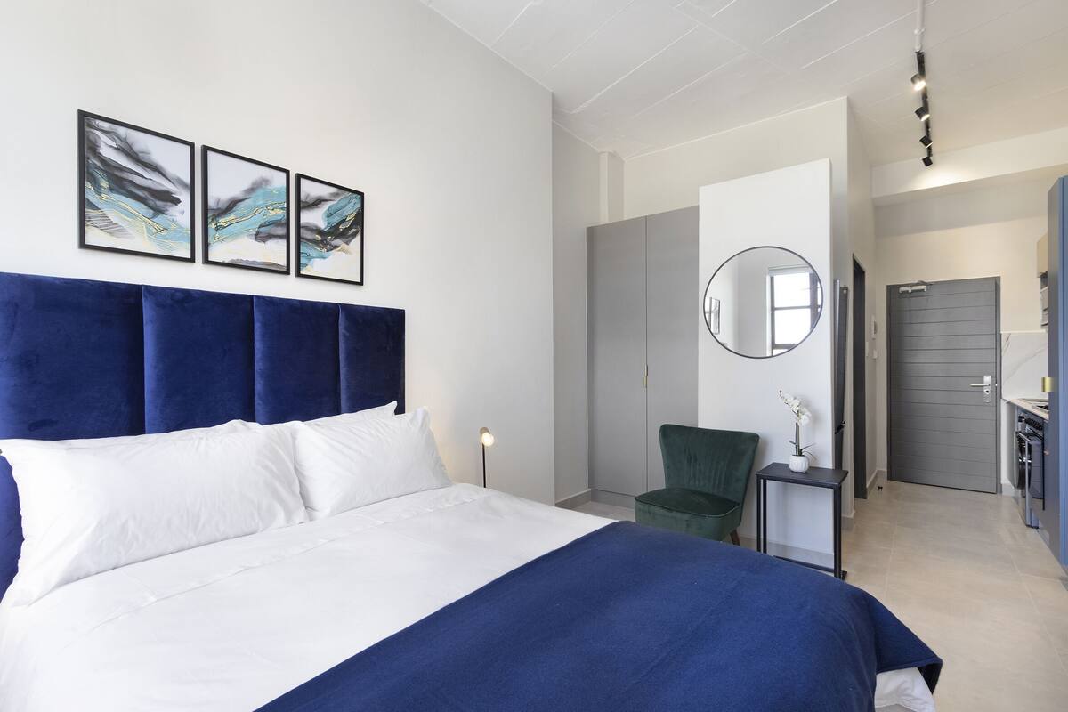 Una habitación de hotel moderna con una cama grande, una silla verde, una mesita con un florero y un espejo en la pared.