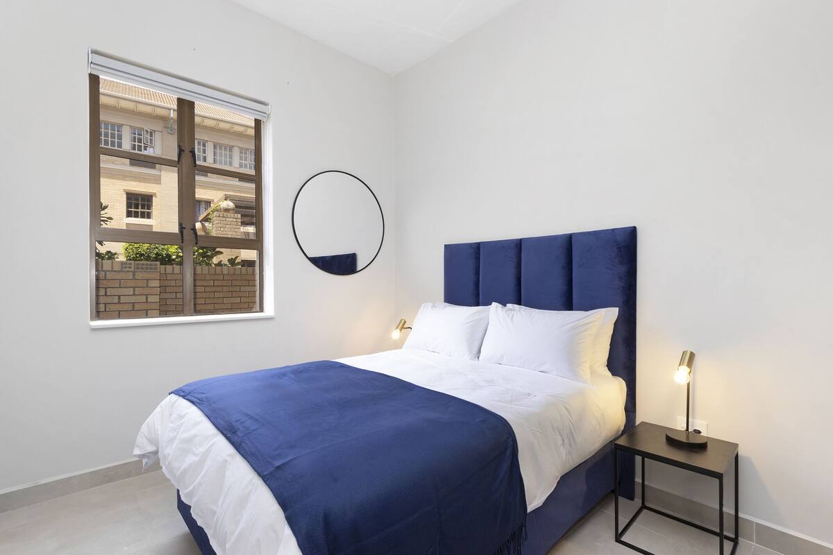 Un dormitorio con una cama, una mesita de noche, una lámpara, una ventana y un espejo.