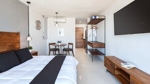 A modern hotel room with a large bed, a desk with chairs, a small table with a plant, a wardrobe, and a TV mounted on the wall. - BoHo lofts & studios new modern w/ ocean-view pool (Playa del Carmen)