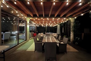 An outdoor dining area with a long table and chairs, surrounded by glass walls and a wooden ceiling with hanging lights. - Chalet with private pool and jacuzzi (Méx.)