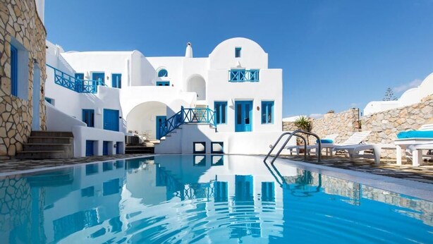 Zona de piscina con edificios blancos y detalles azules, muros de piedra y un cielo azul despejado.