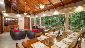 A wooden dining area with a long table set for six, surrounded by chairs. There is a seating area with two sofas and a small bar area in the background. - Casa Bamboo with Guest House | Colonial Estate | Sleeps 14 | Manuel Antonio (Manuel Antonio)