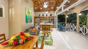 A spacious open-plan living area with a kitchen, dining table with colorful flowers, and a balcony with white railings. - Casa Bamboo with Guest House | Colonial Estate | Sleeps 14 | Manuel Antonio (Manuel Antonio)