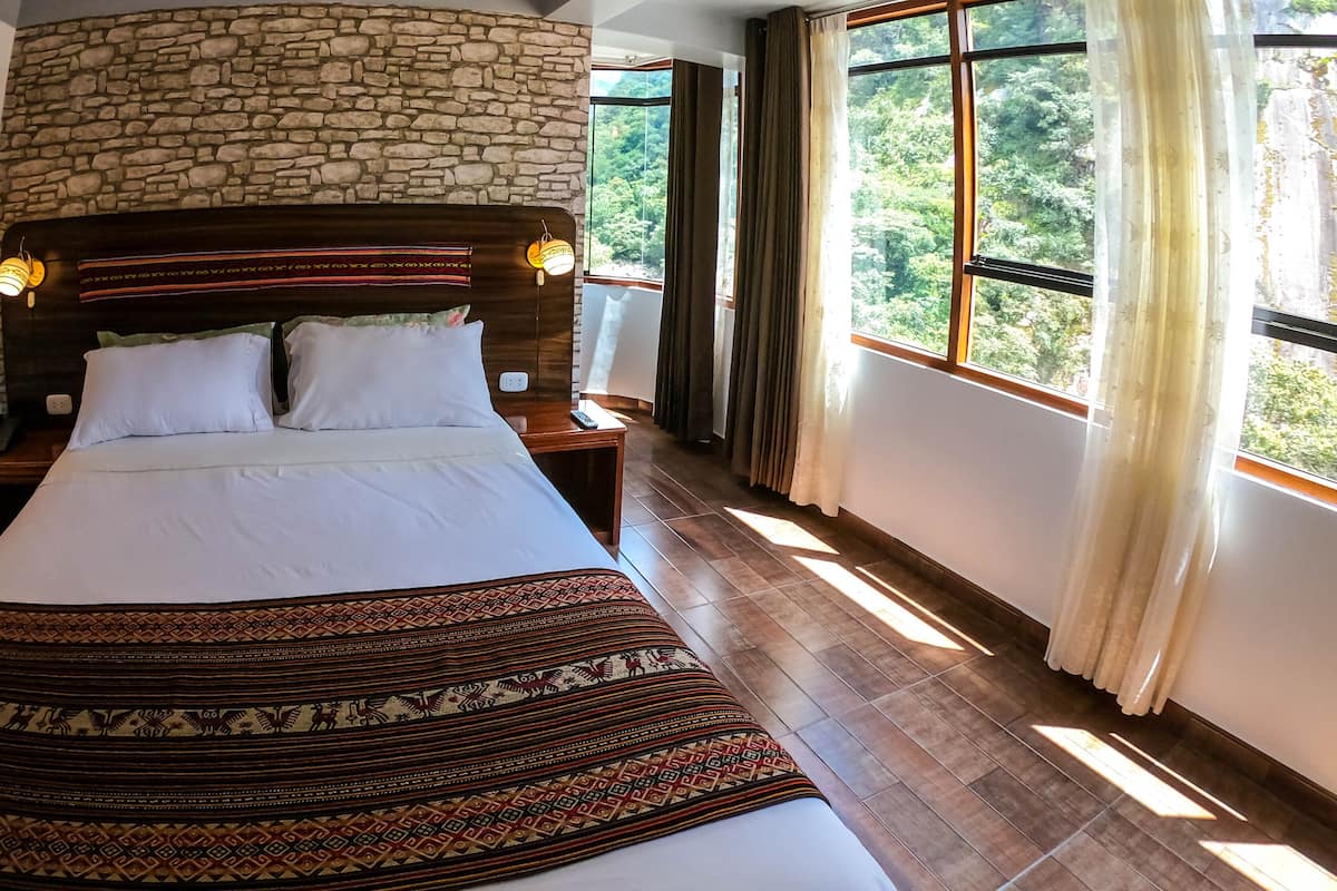 Ein Schlafzimmer mit einer Steinwand, einem Bett mit weißer Bettwäsche, einem gemusterten Teppich und großen Fenstern mit Vorhängen.