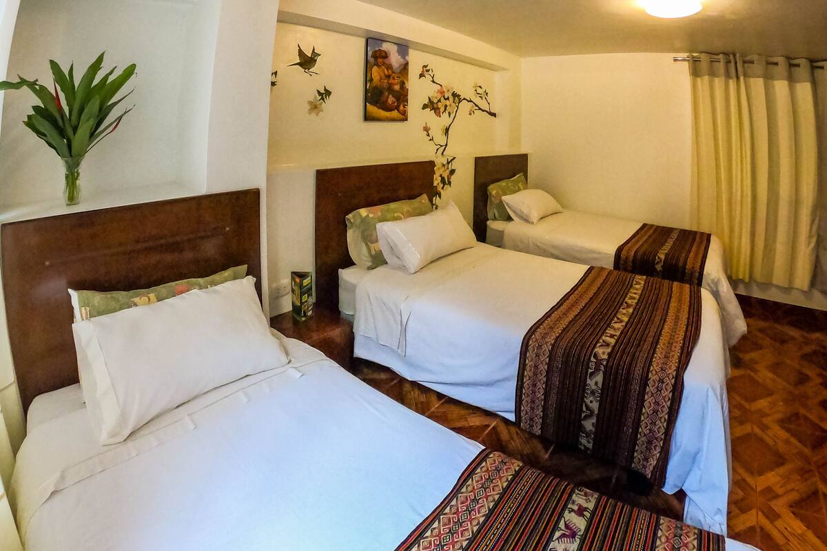 Ein Hotelzimmer mit drei Betten, Holzboden, einem Gemälde an der Wand und einer Pflanze links.