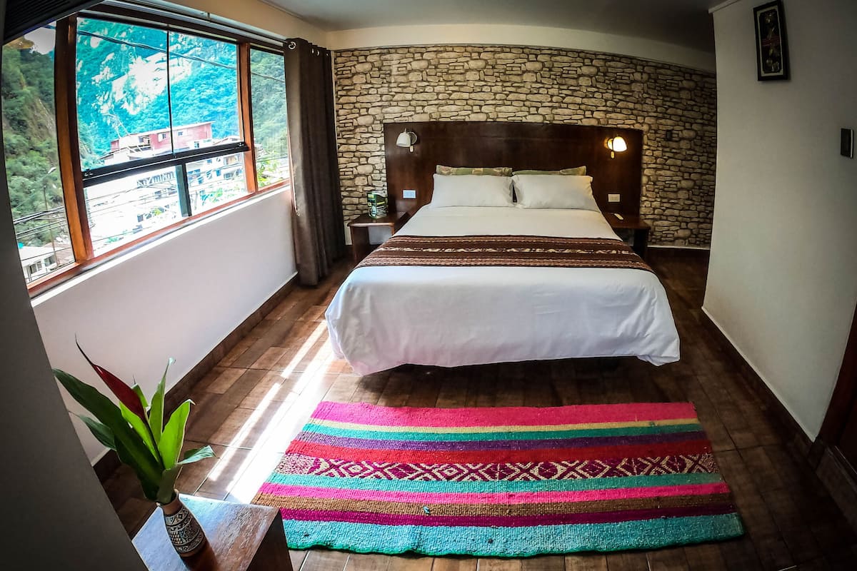 Ein Schlafzimmer mit einer Steinwand, einem Bett mit weißer Bettwäsche, einem farbenfrohen Teppich und einem Fenster mit Blick auf Grünflächen.