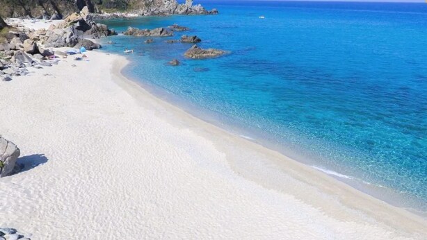 Ein Strand mit klarem, blauem Wasser, weißem Sand und Felsformationen.