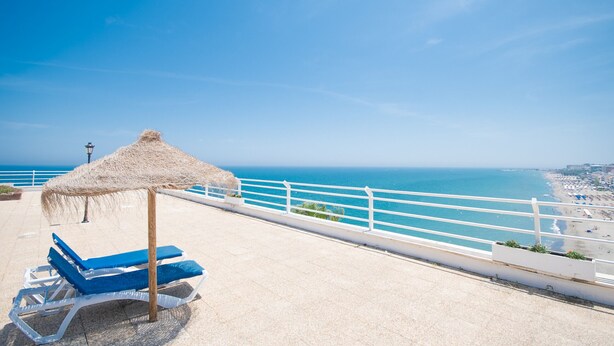 Une terrasse au bord de la plage, dotée d’un parasol en chaume et de chaises longues bleues, offrant une vue sur la mer.