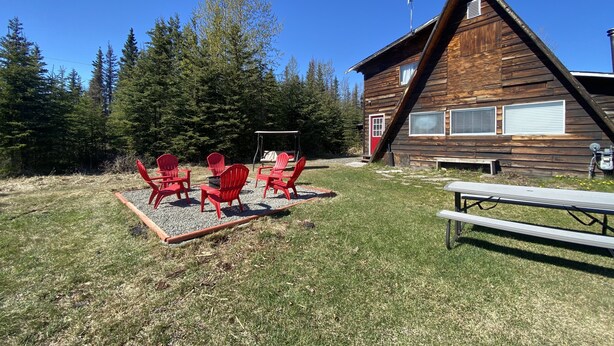 Una cabaña de madera con puerta roja, rodeada de césped, sillas Adirondack rojas y una mesa de picnic.