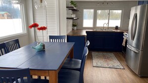 Una cocina moderna con una mesa de comedor de madera, sillas azules, un refrigerador de acero inoxidable y un ventanal con vista a árboles.