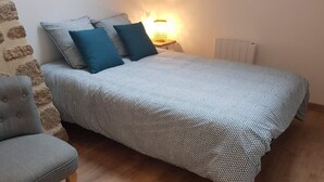 A bedroom with a patterned bedspread, two pillows, a bedside lamp, a radiator, and a grey armchair. - Nature cottage in Suisse Normande (Athis Val de Rouvre)