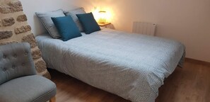 Une chambre à coucher avec une couvre-lit à motifs, deux oreillers, une lampe de chevet, un radiateur et un fauteuil gris.