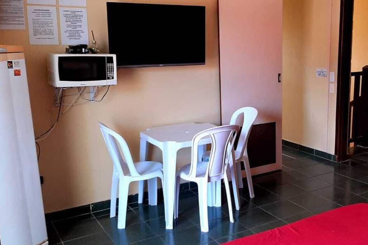 Une petite pièce avec une table et des chaises blanches, un four à micro-ondes, une télévision et un lit superposé avec des coussins rouges.