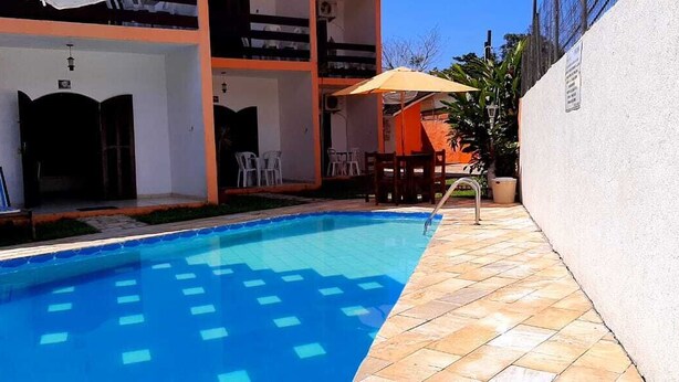 A swimming pool with a tiled deck, outdoor seating, and a covered area with chairs and an umbrella. - Pousada Villa Garden (São Sebastião)