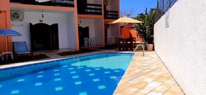 A swimming pool with a tiled deck, outdoor seating, and a covered area with chairs and an umbrella. - Pousada Villa Garden (São Sebastião)