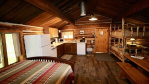 A rustic cabin with a wooden interior, a kitchen with basic appliances, a bed with a striped blanket, and a bunk bed area. - Gold Camp - A Classy-Rustic Gem (Clark)