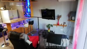 A compact living space with a sofa, a small table, a TV mounted on the wall, and a bar area with bottles and glasses.