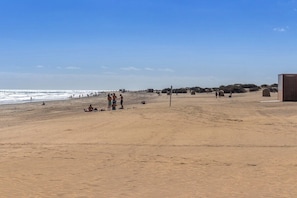 Praia arenosa e extensa com pessoas e tendas, sob um céu claro.