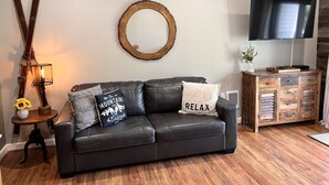 A living room with a black leather sofa, a wooden cabinet, a TV, and decorative items. - B213 - Loon Mountain Resort Studio with Pool, Hot Tub & Full Kitchen (Lincoln)