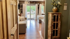 A hotel room with a wooden floor, a sofa with a pillow labeled "RELAX", a wooden cabinet, and a view of the outdoors through a glass door. - B213 - Loon Mountain Resort Studio with Pool, Hot Tub & Full Kitchen (Lincoln)