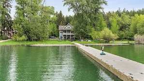 Una propiedad junto al lago con un muelle que se adentra en el agua, rodeada de abundante vegetación y una casa de dos pisos.
