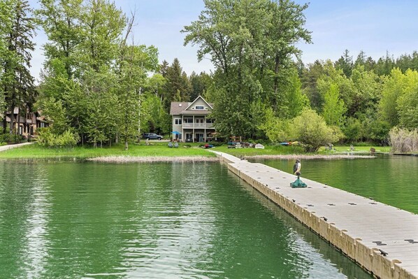 A lakeside property with a dock extending into the water, surrounded by lush greenery and a two-story house. - Waterfront, Whitefish Lake apartment! (Whitefish)