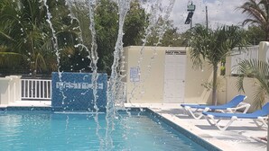 En swimmingpool med springvand, et skilt med teksten "WESTMORNE" og liggestole langs poolkanten.