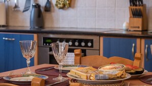 Een eettafel gedekt met borden, glazen en etenswaren in een keuken met blauwe kasten en diverse keukengerei.