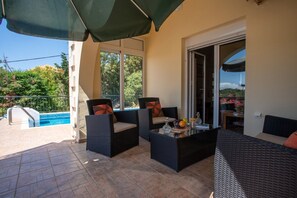 En terrasse med to fletstole, et bord med frugt og stearinlys, og en swimmingpool i baggrunden.