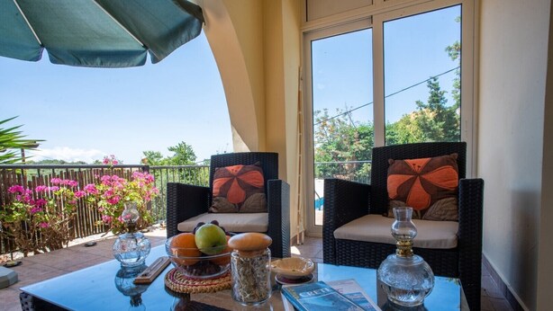 Een balkon met rieten stoelen, een glazen tafel en uitzicht op roze bloemen en groen.