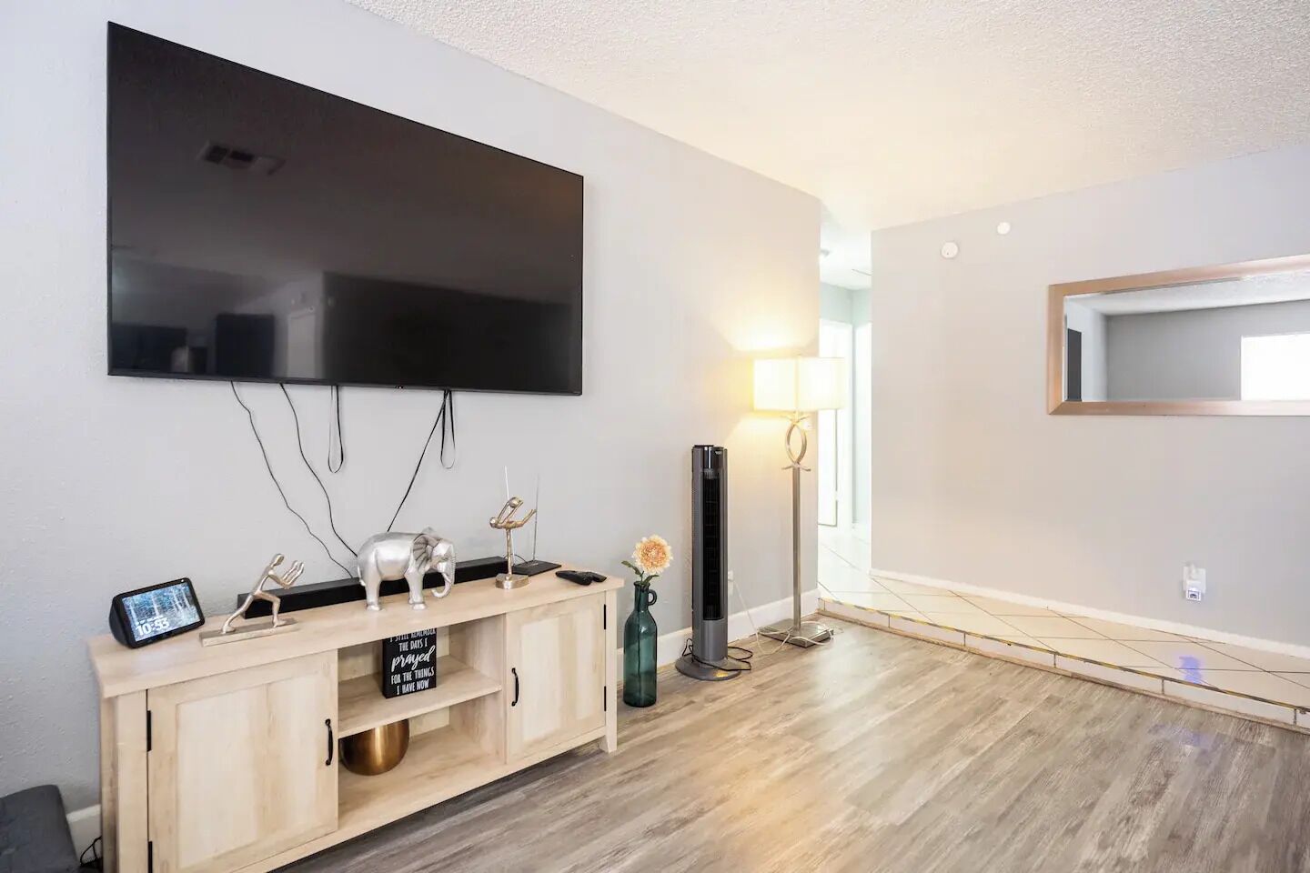 Ein modernes Wohnzimmer mit einem wandmontierten Flachbildfernseher, einem Holz-Konsolentisch mit Dekorationen, einer Stehlampe und einem großen Spiegel.