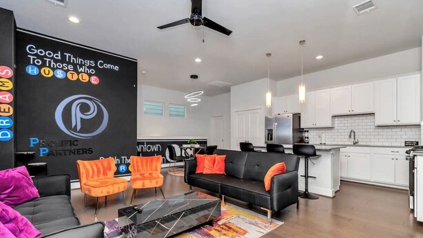 A modern living room with a black sofa, orange chairs, and a black coffee table. There is a kitchen area with white cabinets and a stainless steel refrigerator. - Stylish Townhouse in Central Houston (Houston)