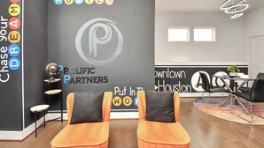 A modern hotel lobby with a black wall featuring motivational text and a logo. There are two orange chairs in the foreground, a round table with chairs, and a decorative globe on a stand. - Stylish Townhouse in Central Houston (Houston)