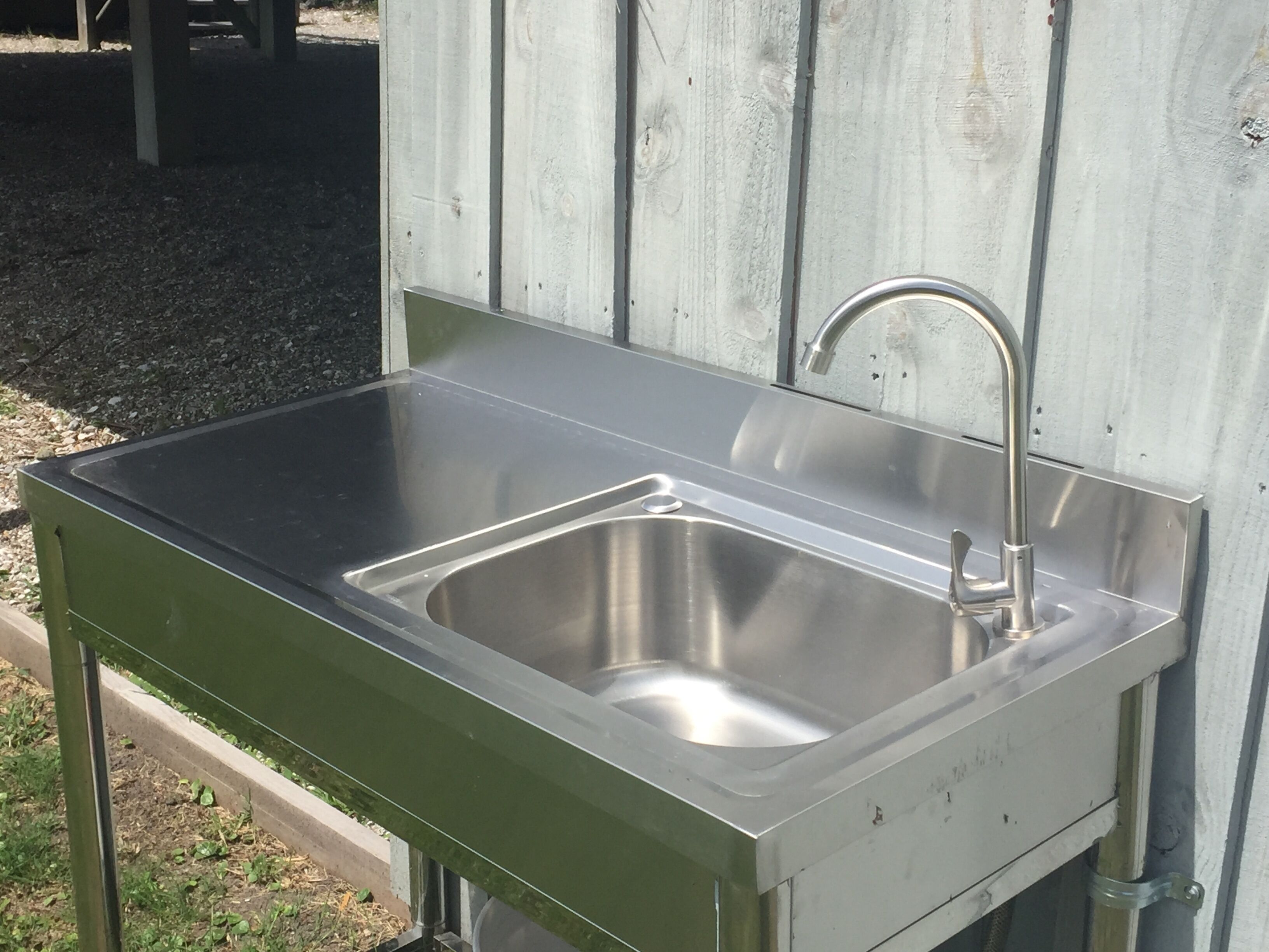 A stainless steel outdoor sink with a single faucet, mounted on a green metal frame, against a wooden wall.