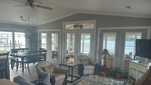 A spacious living room with a large sofa, a coffee table, and a television. There are several chairs and a dining table. The room has large windows with a view of the water. - Rivendock Rookery II: Waterfront Home w/Dock & Stunning Views (Oriental)